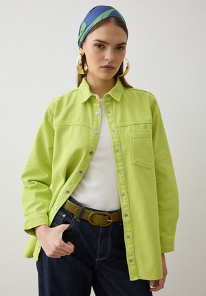 Jeune femme portant une chemise boutonnée vert lime, un haut blanc, un jean foncé, un foulard bleu et vert sur la tête, et de grandes boucles d'oreilles dorées en anneaux.