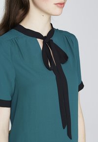Blusa turchese con colletto nero a laccetti, maniche corte e scollatura a chiave. Tessuto morbido, leggero e con leggeri arricciamenti sulle spalle.