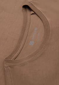 Braunes Baumwoll-T-Shirt mit Rundhalsausschnitt, sichtbaren Nähten und einem Etikett mit Marken- und Größeninformationen.