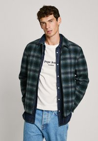 Žalios ir jūros mėlynos plaid flanelinės marškiniai su džinsiniu apykakle, dėvimi virš baltos marškinėlės su juodu tekstu, derinami su mėlynais džinsais.