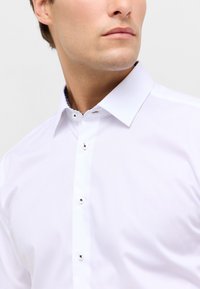 Witte button-up shirt met een gespreide kraag, zwarte knoopaccenten en contrasterende textuurdetails op de binnenkraag. Glad materiaal.