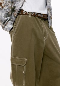 Persona che indossa pantaloni cargo oliva con cuciture bianche, una cintura marrone con grandi occhielli metallici, una camicia bianca e un giubbotto a fantasia.