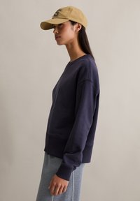 Granatowa bluza dresowa o luźnym kroju, z ściągaczami na rękawach i dole; w połączeniu z jasnoszarymi spodniami. Beżowa czapka z literowym zdobieniem. Prosta faktura.