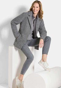 Femme souriante portant un manteau gris, un pull et un pantalon, assise avec une jambe posée sur un piédestal cylindrique blanc et les mains dans les poches.
