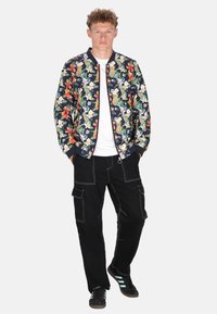 Giubbotto bomber floreale in blu navy con fiori colorati, abbinato a pantaloni cargo neri con diverse tasche e cuciture bianche.