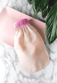 Beige Stoffbeutel mit einem rosa Gummiband, auf dem der Text "pure" steht. Glatte Textur, abgerundeter Boden, auf einem rosa Teller mit Marmorhintergrund platziert.