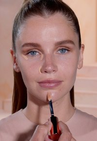 Vloeibare concealer in een applicator wordt op het gezicht van een model aangebracht. De concealer is lichtbeige en het model heeft een heldere huid en lichte make-up.