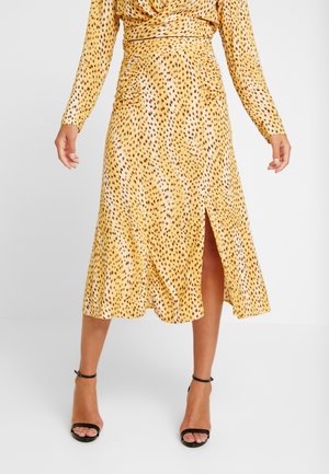 Femme portant une jupe midi à imprimé léopard jaune avec une fente à l'avant, associée à des escarpins noirs à bride de cheville et un haut à manches longues assorti.