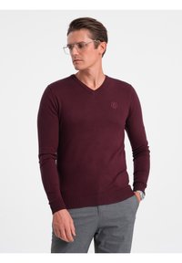 Maglione bordeaux a scollo a V, tessuto lavorato a maglia, design aderente, polsini e orlo a coste, con un logo discreto sul petto, indossato con pantaloni grigi.
