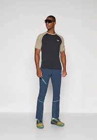 Hombre vestido con una camisa de manga corta negra y beige, pantalones deportivos azul marino con acentos azules, zapatillas de correr verde y azul, y gafas de sol negras.