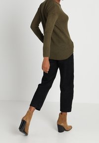 Pull vert olive en tricot à manches longues, associé à un pantalon en velours côtelé noir et des bottines couleur beige à talon épais noir.
