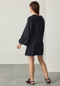 Denimkjole i mørkeblå med Peter Pan-krave, lange pufærmer og en let udvidet silhuet, kombineret med mørkebrune slip-on sko.