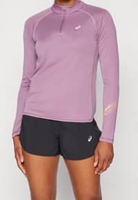 Naisvartalo, jolla on yllään mauvenvärinen pitkähihaoinen urheilupaita, jossa on lyhyt vetoketju, sekä mustat juoksushortsit, joissa molemmissa on valkoiset Asics-logot.