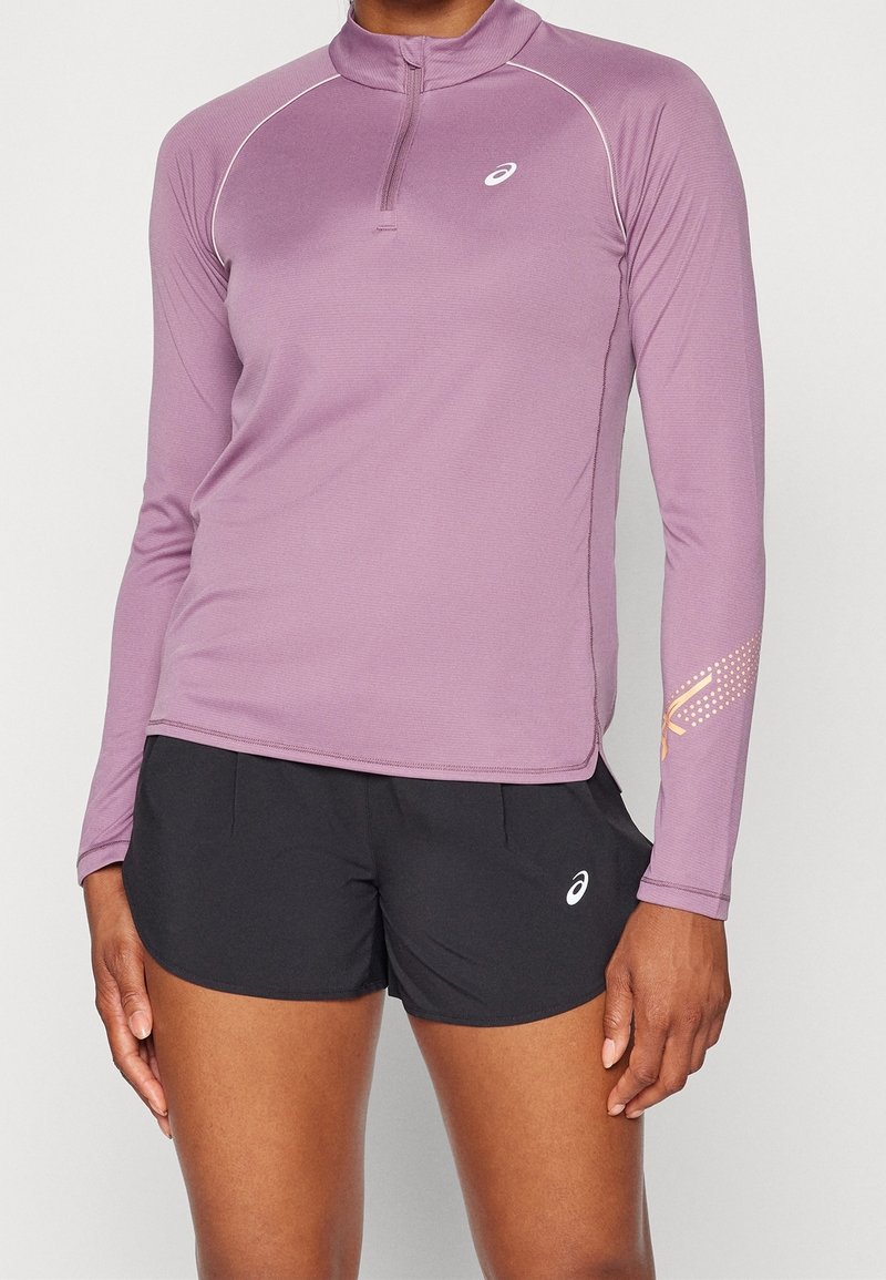 Naisvartalo, jolla on yllään mauvenvärinen pitkähihaoinen urheilupaita, jossa on lyhyt vetoketju, sekä mustat juoksushortsit, joissa molemmissa on valkoiset Asics-logot.