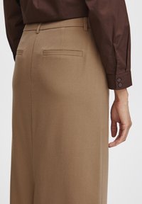 Bruine broek met een getailleerde fit, voorzien van twee achterzakken en een gladde, geweven textuur. De tailleband zit iets boven de heupen.