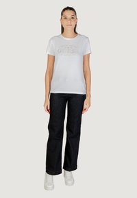 T-shirt bianca a maniche corte con logo metallico "GUESS Los Angeles", abbinata a jeans in denim scuro e sneaker platform bianche.
