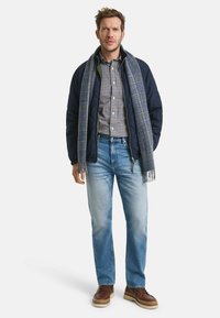 Marineblauwe gewatteerde jas, geblokt shirt, blauwe jeans, grijze sjaal met ruitpatroon, bruine schoenen met lichte zolen; algehele casual look.