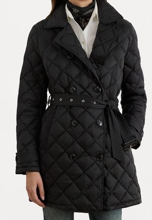 Kvinde iført en sort quiltet dobbeltradet frakke med bælte i taljen, bred krave, manchetstropper og en hvid skjorte med et sort tørklæde.