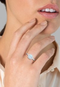 Bague en argent avec une pierre centrale ronde de couleur bleu clair entourée de petites pierres claires, montées sur une bande texturée. Main touchant délicatement le dessus.