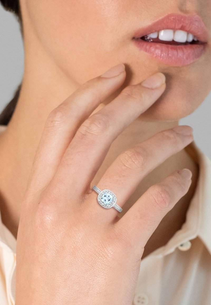 Bague en argent avec une pierre centrale ronde de couleur bleu clair entourée de petites pierres claires, montées sur une bande texturée. Main touchant délicatement le dessus.