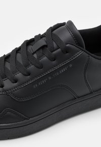 Baskets en cuir noir avec lacets plats noirs, surface texturée et coordonnées géographiques embossées sur le côté. Semelle en caoutchouc profilée basse.