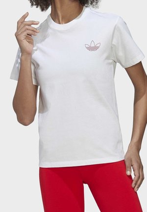 Personne portant un t-shirt blanc Adidas avec un petit logo sur la poitrine et des leggings rouge vif, posant avec une main près du visage.