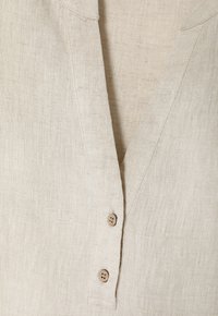 Detailní záběr béžové lněné košile s výstřihem do V a dvěma malými kulatými dřevěnými knoflíky na légě.