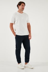 Vīriešu ikdienas apģērbs, kurā iekļauts balts kokvilnas T-krekls ar nelielu logo, kombinēts ar tumši ziliem sporta biksēm un baltām sneakers.