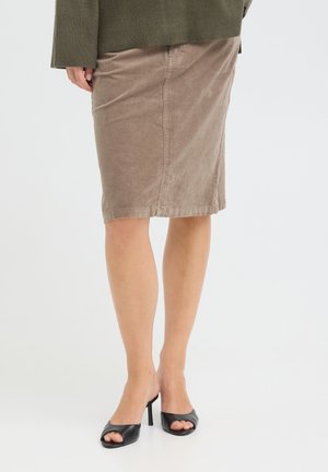 Femme portant une jupe en velours côtelé beige longueur genou, un pull vert olive et des chaussures à talons hauts noires à bouts ouverts, debout sur un sol blanc.