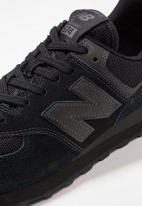 Černé tenisky New Balance 574, vyrobené z kůže a síťoviny, s tkaničkami a výrazným logem "N" v matném provedení. Plná gumová podrážka.