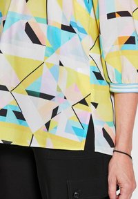 Bunte Bluse mit geometrischem Muster in Gelb, Blau und Schwarz, aus leichtem Stoff und mit kurzen, plissierten Ärmeln.