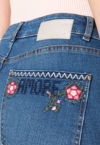 Mörkblå denimjeans med broderad "AMORE" och blommönster på bakfickan; har en ljusläderetikett ovanför fickan.
