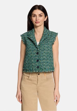Femme portant un gilet sans manches texturé vert-bleu à boutons sur un haut blanc et un pantalon beige taille haute, debout devant un fond uni.