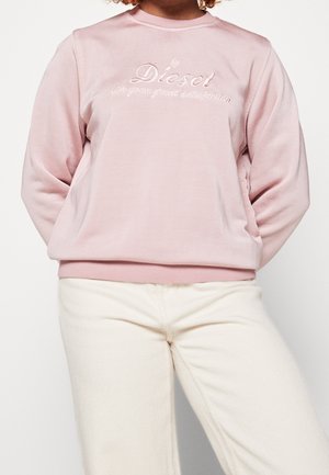Personne portant un sweat-shirt rose clair avec le texte « by Diesel for your great satisfaction » et un pantalon écru, debout les mains derrière le dos.