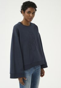 Tumši zila pārspīlēta kapuce ar apaļu kakla izgriezumu, nolaižamām plecu šuvēm un izliekta logotipa dizainu priekšpusē. Mīksta auduma tekstūra.