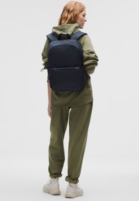 Zaino nero con spallacci imbottiti, forma rettangolare, texture liscia, tasca frontale con zip, indossato con felpa con cappuccio verde oliva e pantaloni da ginnastica.