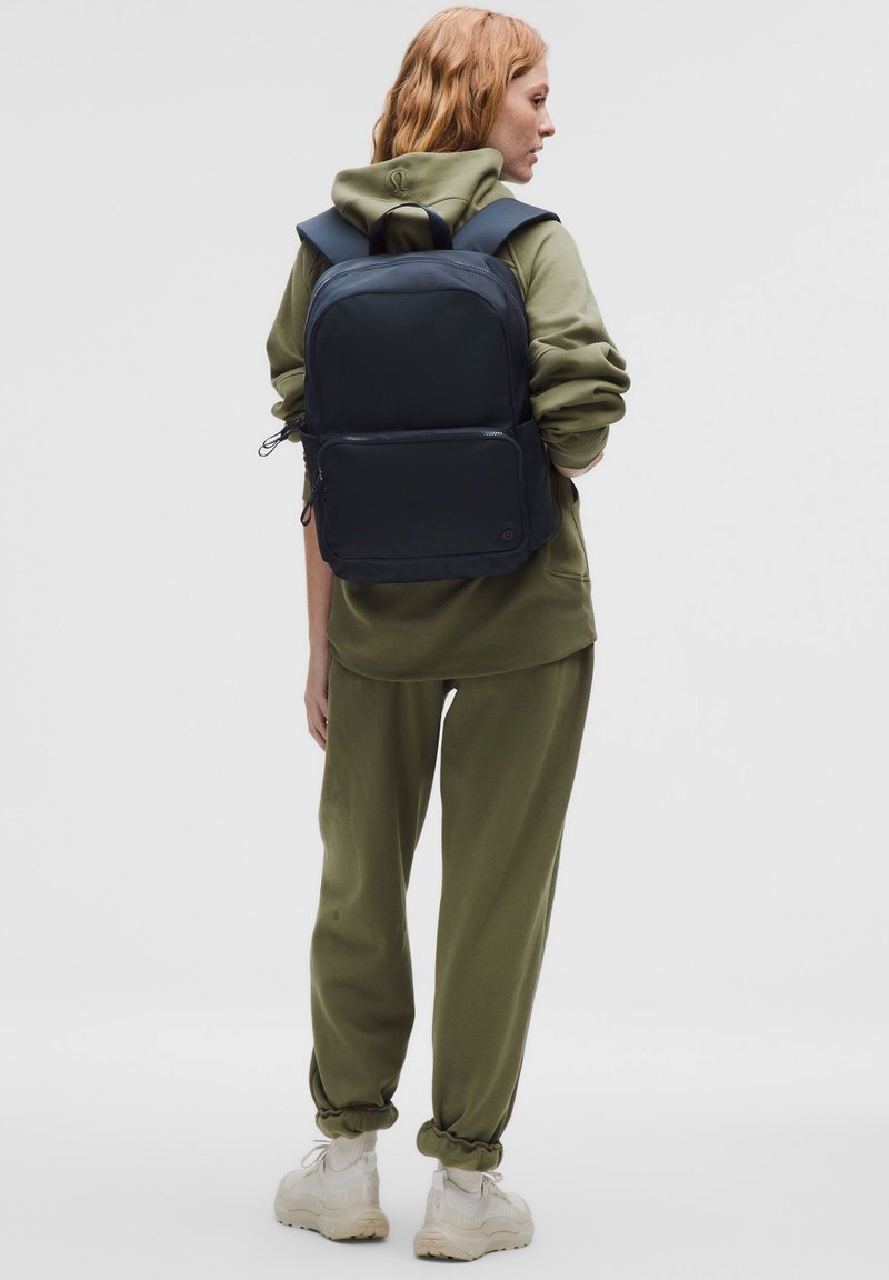 Zaino nero con spallacci imbottiti, forma rettangolare, texture liscia, tasca frontale con zip, indossato con felpa con cappuccio verde oliva e pantaloni da ginnastica.