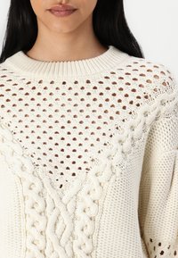 Pull en maille épaisse de couleur crème, présentant un design côtelé texturé et un tissage ouvert au niveau de l'encolure. Le col et les manches ribbés complètent le style.