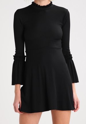 Robe noire à côtes avec un col haut, des manches évasées avec des accents élastiques, et une jupe évasée tombant au-dessus de la mi-cuisse.