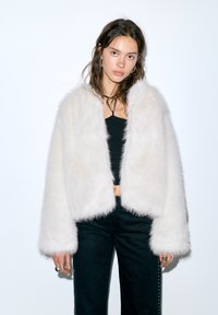Jachetă scurtă din blană artificială albă cu o textură moale, cu mâneci largi și tiv rotunjit, purtată peste un top negru.