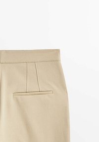 Pantalon beige tailleur avec une poche passepoilée arrière et un détail de ceinture cousu sur fond blanc.