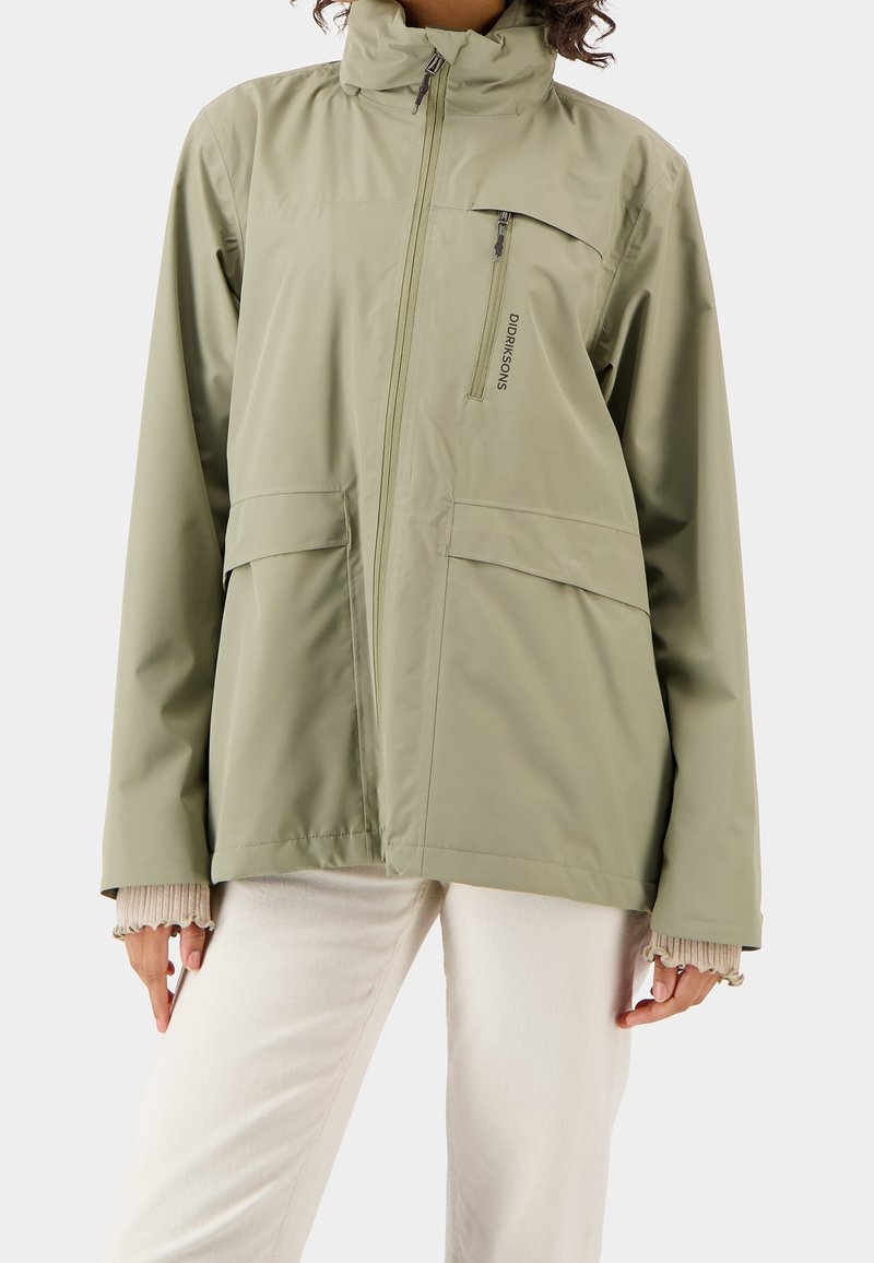 Veste zippée vert olive clair avec col montant, poche poitrine verticale, poches à rabat, portée sur un pantalon blanc et des poignets en maille beige.