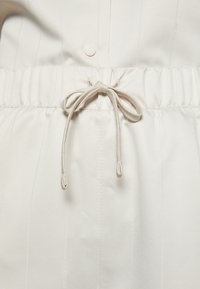 Gros plan d'une ceinture élastique blanche avec un cordon noué et un bouton sur un tissu blanc assorti au-dessus.