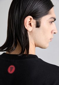 Svart t-skjorte med rund hals, som har et rødt grafisk logo på baksiden. Stoffet ser mykt ut med en glatt tekstur.