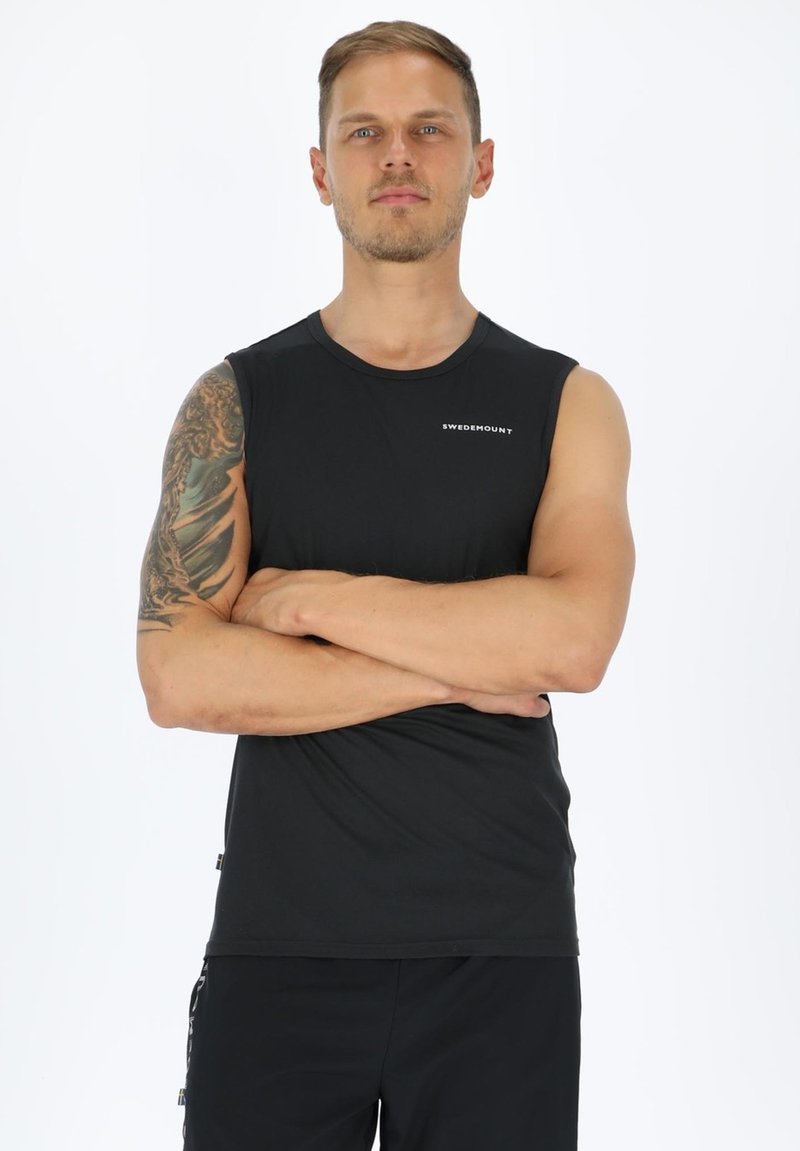 Mann mit tätowiertem linkem Arm trägt schwarzes ärmelloses SWEDEMOUNT-Oberteil und steht mit verschränkten Armen vor weißem Hintergrund.