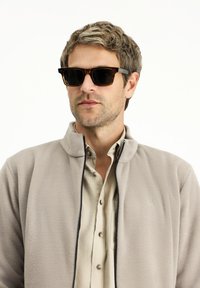 Fleecejacke in Hellgrau, mit Reißverschluss und hohem Kragen. Hellbraunes, knöpfbares Hemd darunter. Dunkle Sonnenbrille mit Schildpattrahmen.