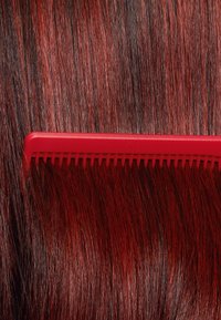 Pettine di plastica rossa appoggiato su lunghi capelli lisci di un rosso sfumato. Il pettine presenta denti fini e un bordo dritto, in contrasto con la texture dei capelli.