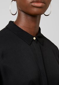 Gros plan d'une personne portant de grandes boucles d’oreilles argentées en forme d’anneau et un haut noir à col avec un seul bouton doré visible.