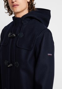 Cappotto in misto lana di colore navy con chiusura a toggles, ampie tasche frontali e cappuccio. Presenta una piccola etichetta con logo sul maniche.
