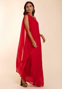 Vestido maxi rojo y fluido de chiffon con un diseño de un solo hombro y un panel tipo capa. Presenta unos zapatos de tacón alto con tiras.